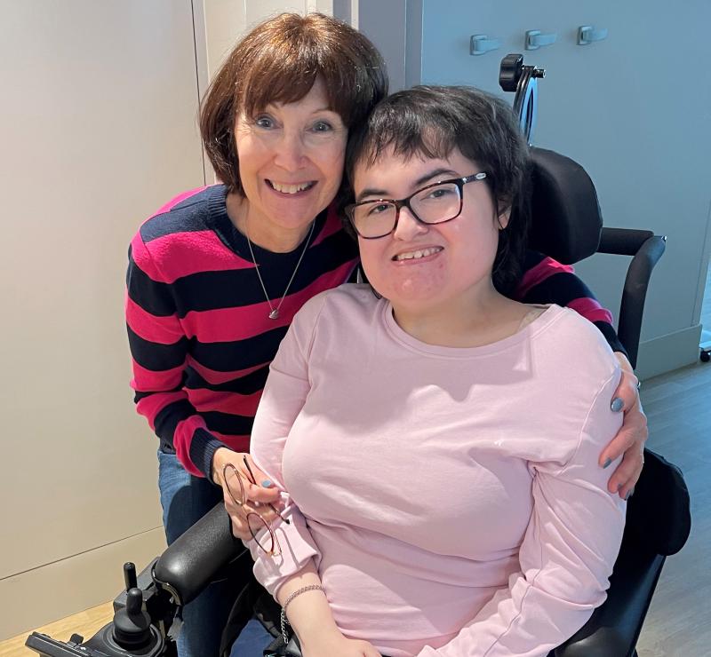 Une femme aux cheveux bruns et courts, portant un pull rayé rose et bleu marine, enlace affectueusement une jeune femme assise dans un fauteuil roulant électrique. Cette dernière porte un haut rose pâle et des lunettes, et sourit doucement. Elles sont proches l'une de l'autre, avec une expression chaleureuse et complice. L’arrière-plan montre un intérieur lumineux et moderne.