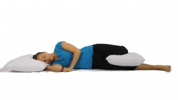 Une femme est allongée sur le côté gauche dans un lit, la tête appuyée sur un oreiller, un second oreiller placé entre ses genoux. Elle adopte une position de sommeil ergonomique, maintenant l’alignement du dos, du bassin et des hanches.