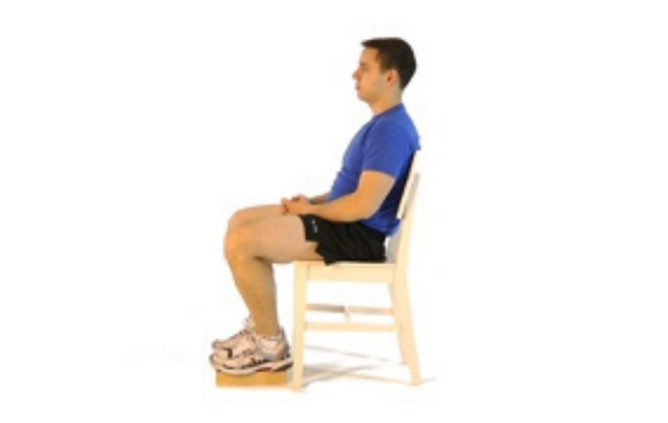 Un homme est assis sur une chaise droite, le dos bien aligné, les épaules détendues et les mains croisées sur les cuisses. Ses pieds sont posés sur un petit bloc pour surélever légèrement les jambes, favorisant une posture assise stable et ergonomique.