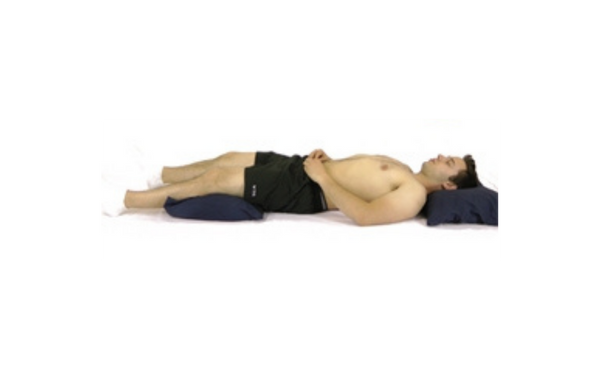 Un homme est allongé sur le dos, les mains posées sur l’abdomen. Sa tête repose sur un oreiller, et un second oreiller est placé sous les genoux pour soutenir la courbure naturelle du bas du dos, favorisant une position de sommeil ergonomique.