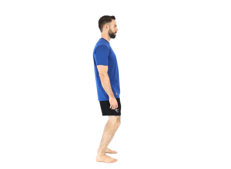 Un homme en t-shirt bleu et short noir se tient debout, pieds nus, les genoux légèrement fléchis, les épaules détendues et le dos droit. Il illustre la posture recommandée pour bien répartir le poids du corps également sur les deux jambes tout en maintenant la courbure naturelle du dos.