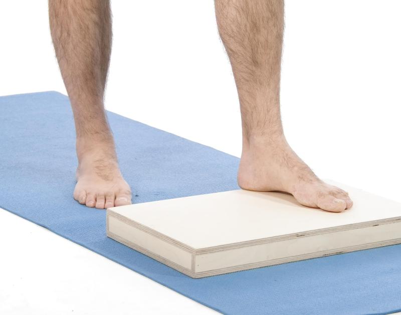 Vue rapprochée de deux pieds nus sur un tapis bleu. Un pied est au sol, l’autre est posé sur une planche de bois d’environ 15 à 20 cm de hauteur, illustrant une technique pour réduire la fatigue en position debout prolongée.