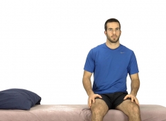 Le même homme est maintenant assis au bord du lit, les mains sur les cuisses, les pieds bien ancrés au sol, prêt à se lever en gardant une posture stable et le dos droit.