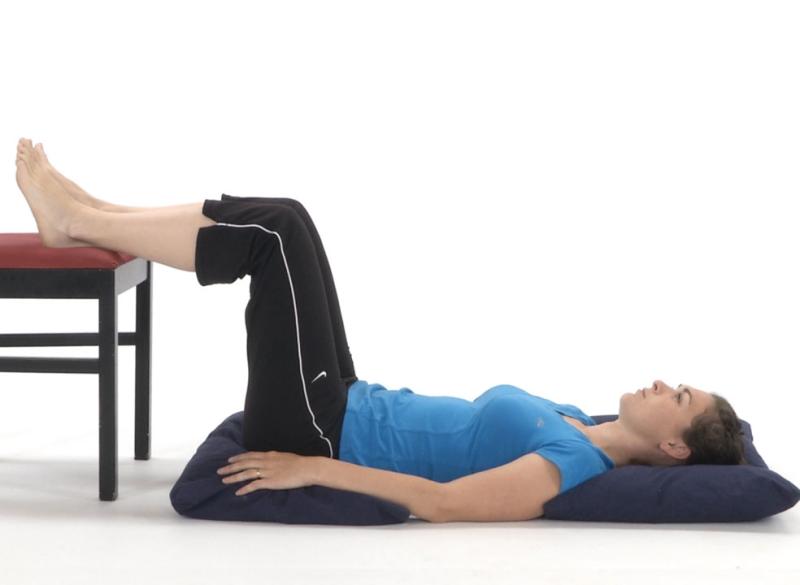 Une femme est couchée sur le dos, les jambes reposant sur une chaise, les genoux pliés à 90 degrés. Sa tête est appuyée sur un oreiller et un coussin soutient son bassin, illustrant une position de décharge pour le bas du dos