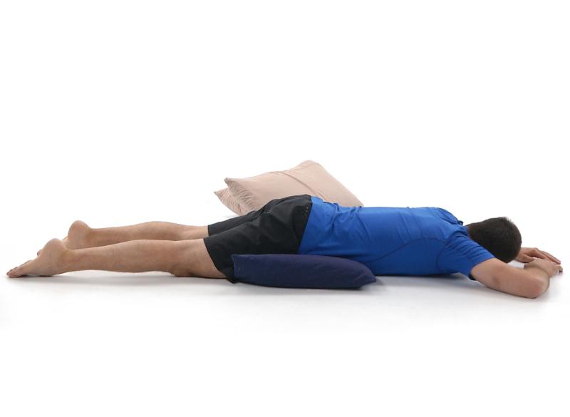 Un homme est couché à plat ventre avec un oreiller sous le bassin et un autre sous la tête. Il repose dans une position de relaxation sur le ventre adaptée pour certaines conditions lombaires.